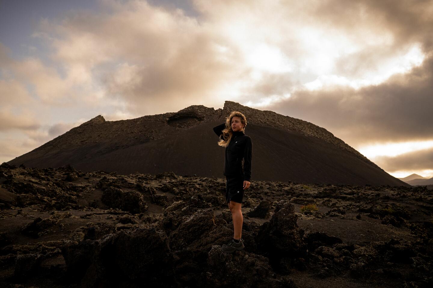 Una mujer rubia de pie, delante de una montaña rocosa, vestida con ropa de Siroko Adventure negra