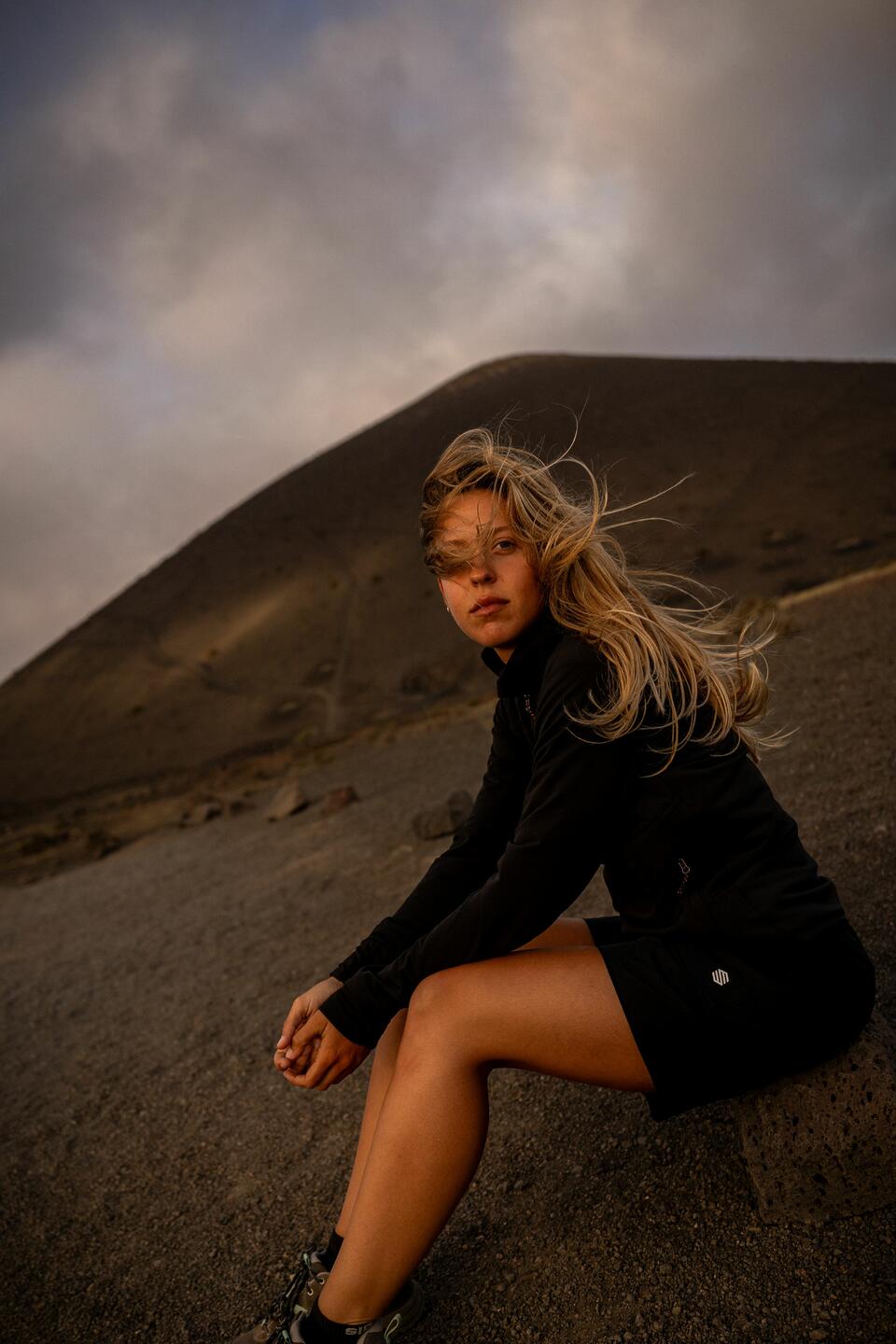 Una mujer rubia sentada, vestida con ropa de Siroko Adventure negra, con una montaña rocosa detrás